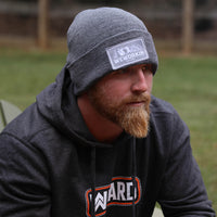 Man pictured from the front/side wearing a We Workin Brand cuffed beanie in Dark Heathered Grey color. Custom 3.5" x 2" patches are sewn on the hats, with the WEWORKIN BRAND text and horizontal half-skull for the design on the patches—woven grey background with white thread for the design and grey merrowed-edge border.