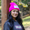 Woman pictured from front wearing a We Workin Brand cuffed beanie in Neon Pink color. Custom 3.5" x 2" patches are sewn on the hats, with the WEWORKIN BRAND text and horizontal half-skull for the design on the patches—woven white background with grey thread for the design and white merrowed-edge border.