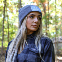 Woman pictured from the side wearing a We Workin Brand cuffed beanie in Dark Heathered Grey color. Custom 3.5" x 2" patches are sewn on the hats, with the WEWORKIN BRAND text and horizontal half-skull for the design on the patches—woven grey background with white thread for the design and grey merrowed-edge border.