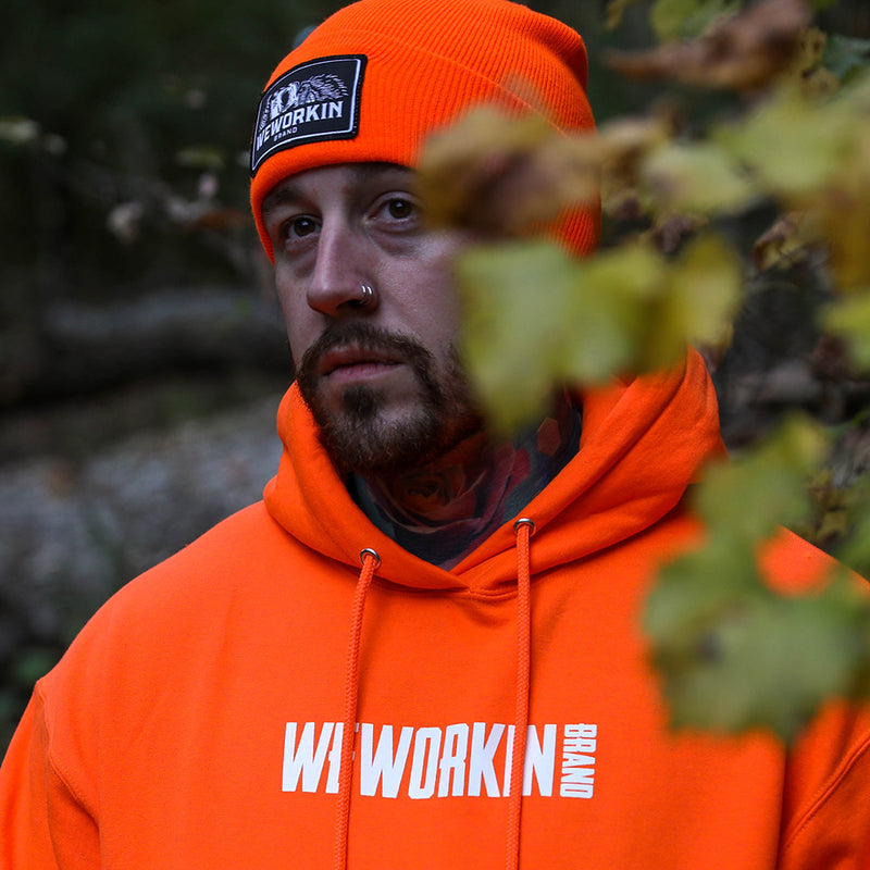 Man pictured from front, in woods, wearing a We Workin Brand cuffed beanie in Neon Safety Orange color. Custom 3.5" x 2" patches are sewn on the hats, with the WEWORKIN BRAND text and horizontal half-skull for the design on the patches—woven black background with white thread for the design and black merrowed-edge border.