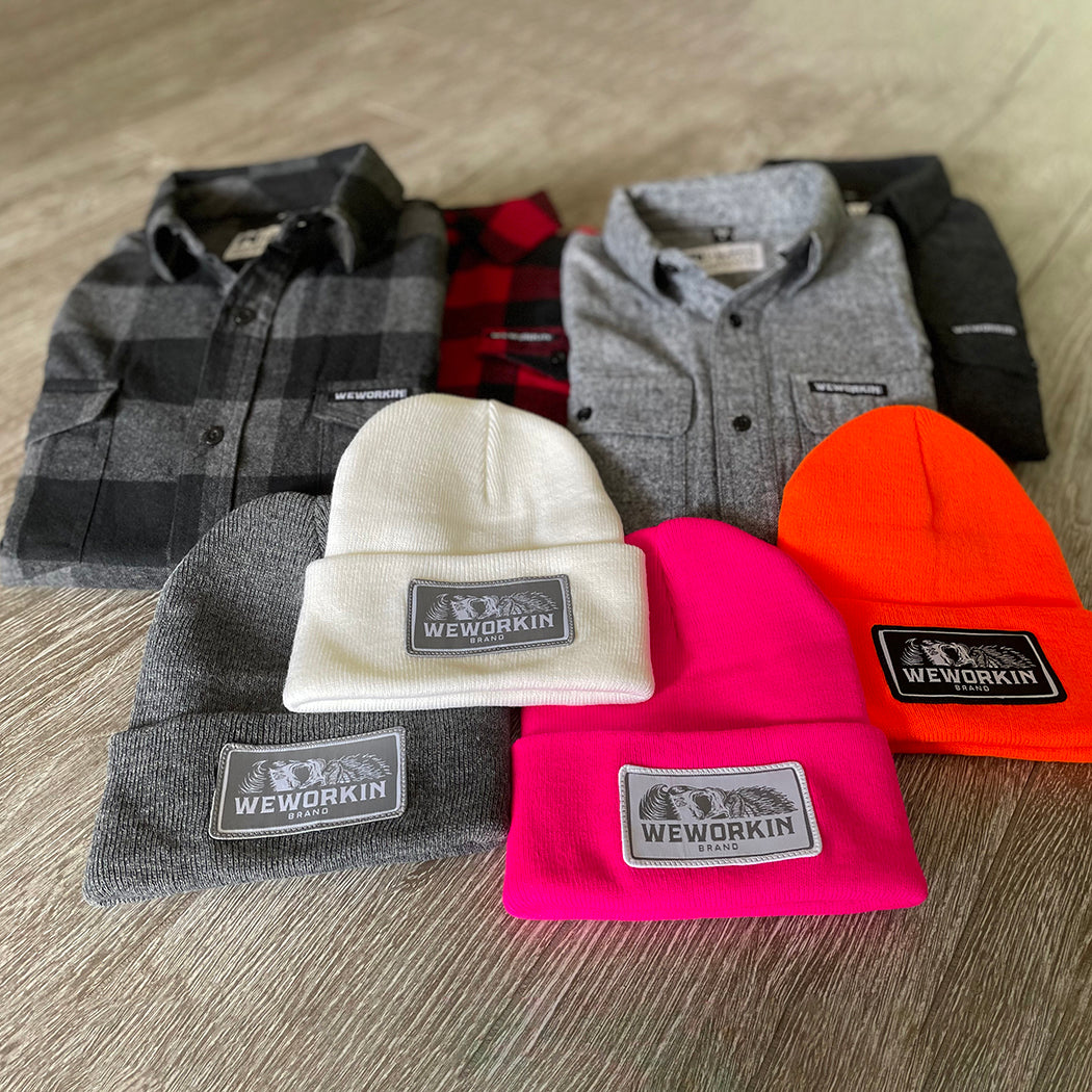 4 We Workin cuffed beanies laying on a grey tile background—4 different colors shown...White, Neon Pink, Neon Safety Orange and Dark Heathered Grey. Custom 3.5" x 2" patches are sewn on the hats, all patch designs have the WEWORKIN BRAND text and horizontal half-skull on the patches—either black, grey or white background with different thread combinations for the design and merrowed-edge borders. 4 We Workin flannels also pictured.