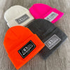 4 We Workin cuffed beanies laying on a grey tile background—4 different colors shown...White, Neon Pink, Neon Safety Orange and Dark Heathered Grey. Custom 3.5" x 2" patches are sewn on the hats, all patch designs have the WEWORKIN BRAND text and horizontal half-skull on the patches—either black, grey or white background with different thread combinations for the design and merrowed-edge borders.