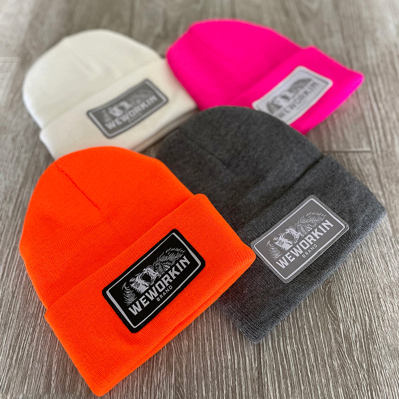 4 We Workin cuffed beanies laying on a grey tile background—4 different colors shown...White, Neon Pink, Neon Safety Orange and Dark Heathered Grey. Custom 3.5" x 2" patches are sewn on the hats, all patch designs have the WEWORKIN BRAND text and horizontal half-skull on the patches—either black, grey or white background with different thread combinations for the design and merrowed-edge borders.
