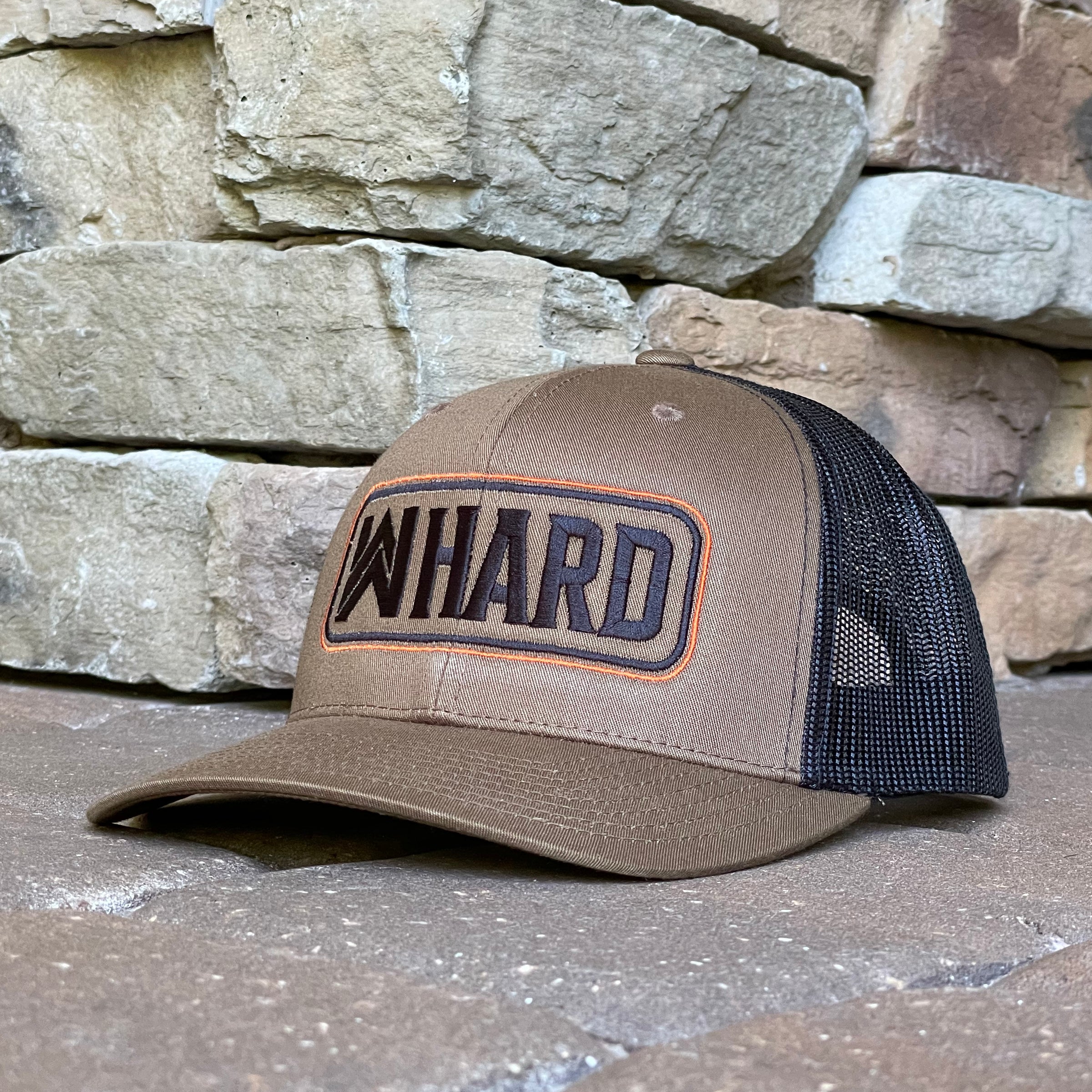 Front left view of a Coyote Brown and Black Retro Trucker hat. Embroidered with WW HARD "patch" style across the front (WW HARD icon/text in black thread, outer outline in orange thread, inner outline in black thread). Front 2 panels and bill are coyote brown, side/back mesh panels are black. Pictured on a brown stone/paver background.