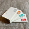 Four Stacks of the WW Clear Stickers 5"w x 3"h ("EVERYBODY WANT$ THE MONEY, NOBODY WANTS THE" words in white letters—"WORK" word highlighted in Grey, Orange, Teal and Red). Each color stack is stacked/fanned out on a grey tile floor.
