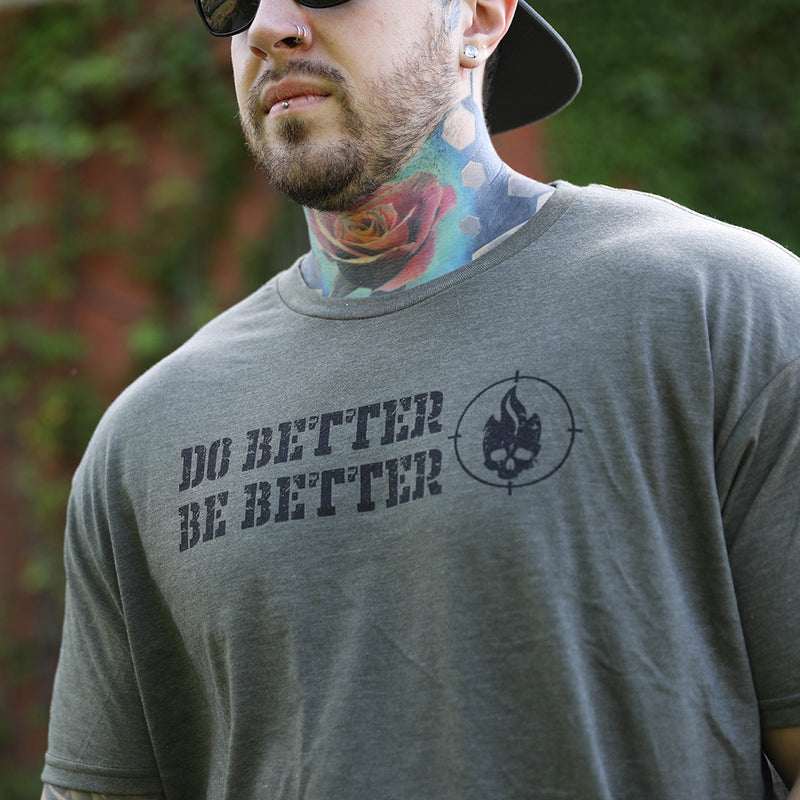 Man pictured from front wearing a WeWorkin heather military green tee—"DO BETTER BE BETTER" text stacked on the left, beside a flaming skull head encircled with a scope mark. He is also wearing a WW Charcoal-Black 7-panel Trucker Hat backwards.