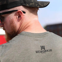 Man pictured from back wearing a We Workin graphic tee in Heather Military Green color. Short sleeve shirt imprinted on back with WeWorkin Brand icon—WW icon centered over top of WEWORKIN BRAND stacked words (printed in black ink). Man is also wearing a WW Black Camo Retro Hat backwards.