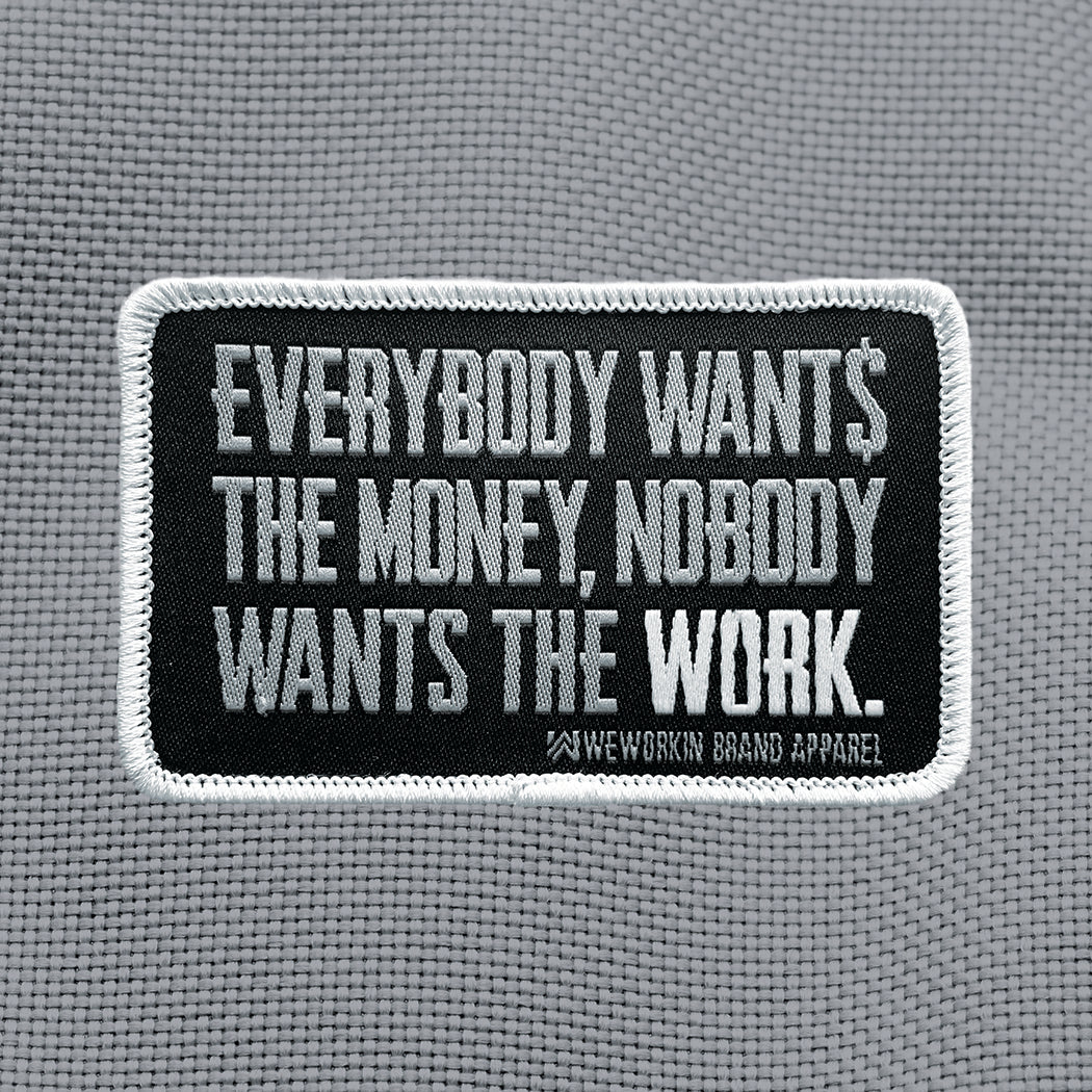 "EVERYBODY WANT$ THE MONEY, NOBODY WANTS THE WORK." text centered large on a velcro-backed patch (both the hook and loop sides provided). [2] thread colors for the design (medium grey and white) on a black woven background, with white merrowed border. 3.5" wide Woven patch displayed on a grey canvas background.