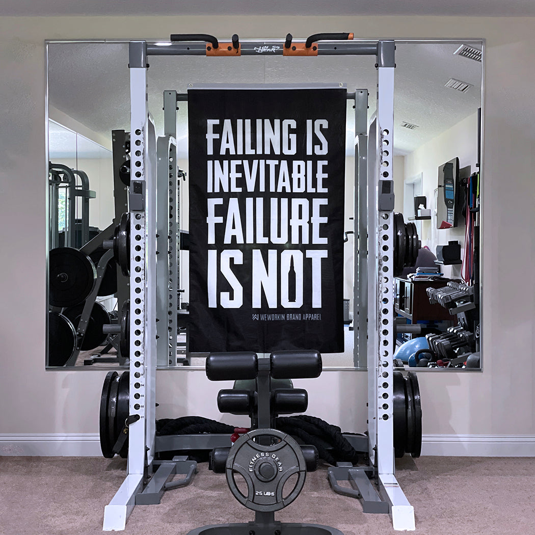 WeWorkin Brand "FAILING IS INEVITABLE—FAILURE IS NOT" flag displayed in a home gym. Each flag measures approximately 3'W x 5'H. The flag has a black background with all white letters. White, double-stitched, thicker top edge for durability, (2) gold grommets (one at top left and one at top right corners).