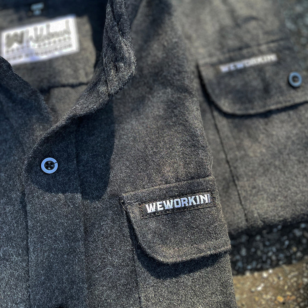 2 We Workin Brand long sleeve Flannel buttoned shirts in charcoal grey are folded and placed on an asphalt, outdoor background. A close-up view of the small/rectangular black with white thread WEWORKIN BRAND woven tag is sewn on the left pocket flap (and a white with black thread WW logo block-tag is sewn on the back panel just below the collar and size tag.)