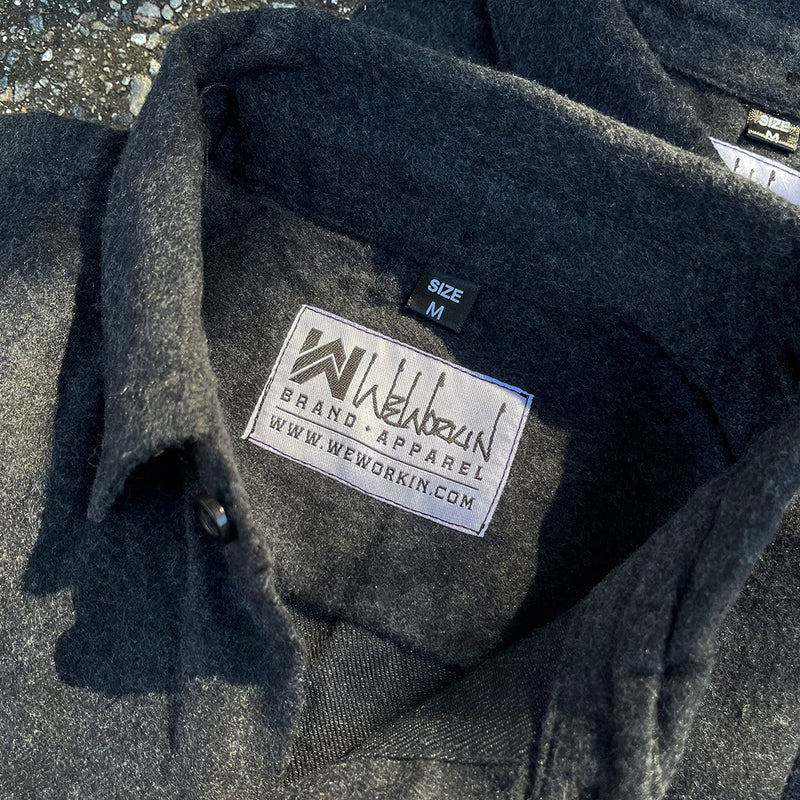2 We Workin Brand long sleeve Flannel buttoned shirts in charcoal grey are folded and placed on an asphalt, outdoor background. A close-up view of the white with black thread WW logo block-tag is sewn on the back panel just below the collar and size tag.