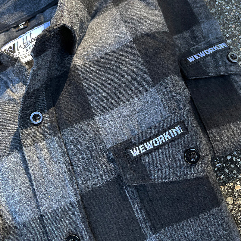 A close up picture of a We Workin long sleeve Flannel buttoned shirt in Grey/Black Buffalo Plaid. A small/rectangular black with white thread WEWORKIN BRAND woven tag is sewn on the left pocket flap. 