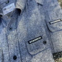 2 We Workin Brand long sleeve Flannel buttoned shirts in Light Heather Grey are folded and placed on an asphalt, outdoor background. A close-up view of the small/rectangular black with white thread WEWORKIN BRAND woven tag is sewn on the left pocket flap (and a white with black thread WW logo block-tag is sewn on the back panel just below the collar and size tag.)