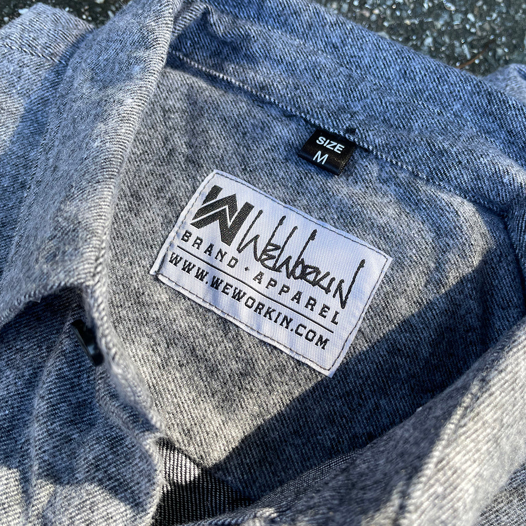 2 We Workin Brand long sleeve Flannel buttoned shirts in Light Heather Grey are folded and placed on an asphalt, outdoor background. A close-up view of the white with black thread WW logo block-tag is sewn on the back panel just below the collar and size tag.