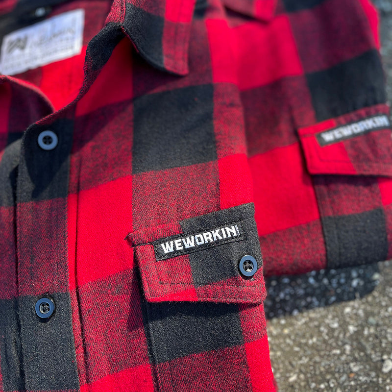 2 We Workin Brand long sleeve Flannel buttoned shirts in Red/Black Buffalo Plaid, folded and placed on asphalt, outdoor background. A close-up view of the small/rectangular black with white thread WEWORKIN BRAND woven tag is sewn on the left pocket flap (and a white with black thread WW logo block-tag is sewn on the back panel just below the collar and size tag.)