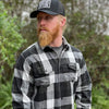 Man pictured from front wearing a We Workin long sleeve Flannel buttoned shirt in Lt Grey/Black Plaid. A small/rectangular black with white thread WEWORKIN BRAND woven tag is sewn on the left pocket flap. Also wearing a WW "Everybody Want$ the Money, Nobody Wants the Work" LG Patch, black snapback Hat.