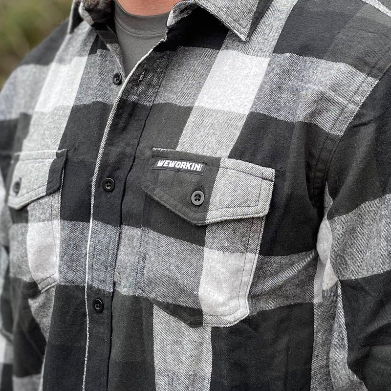 Man pictured from front, close up, wearing a We Workin long sleeve Flannel buttoned shirt in Lt Grey/Black Plaid. A small/rectangular black with white thread WEWORKIN BRAND woven tag is sewn on the left pocket flap. 