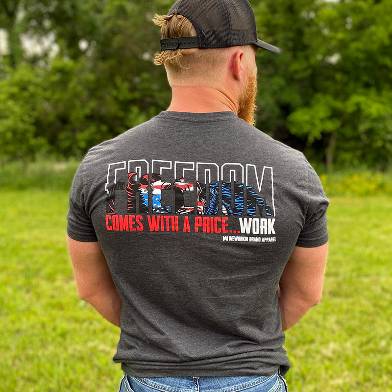 Man outdoors wearing the WE WORKIN men's short sleeved t-shirt in Heather Charcoal Grey. The word FREEDOM outlined in white, with a Red, White, Blue Patriotic (full color) WWB half skull inside the bottom of the letters—"COMES WITH A PRICE...WORK" text is printed below in Red and White ink. All displayed on full upper back width (WEWORKIN BRAND APPAREL and Icon printed small and lower right just below the full back imprint.)