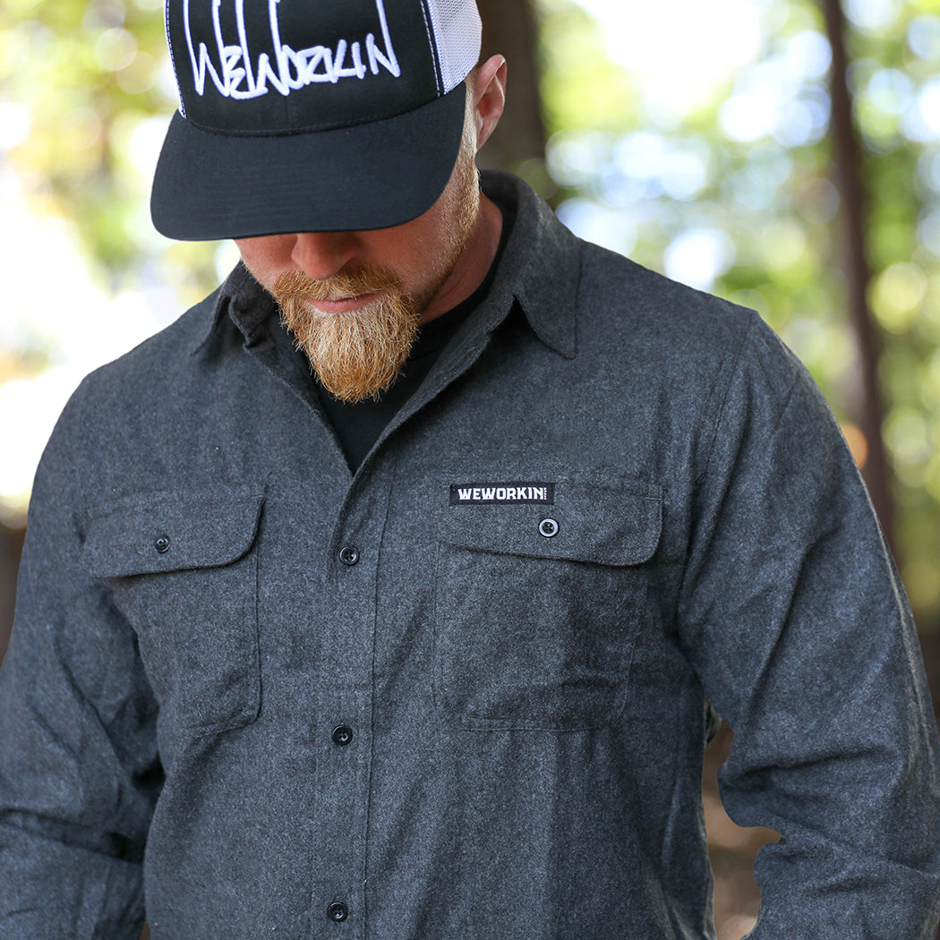 Man pictured from front wearing a We Workin long sleeve Flannel buttoned shirt in charcoal grey, wooded background. A small/rectangular black with white thread WEWORKIN BRAND woven tag is sewn on the left pocket flap. He is also wearing a We Workin Script Retro Trucker hat in Black/White.