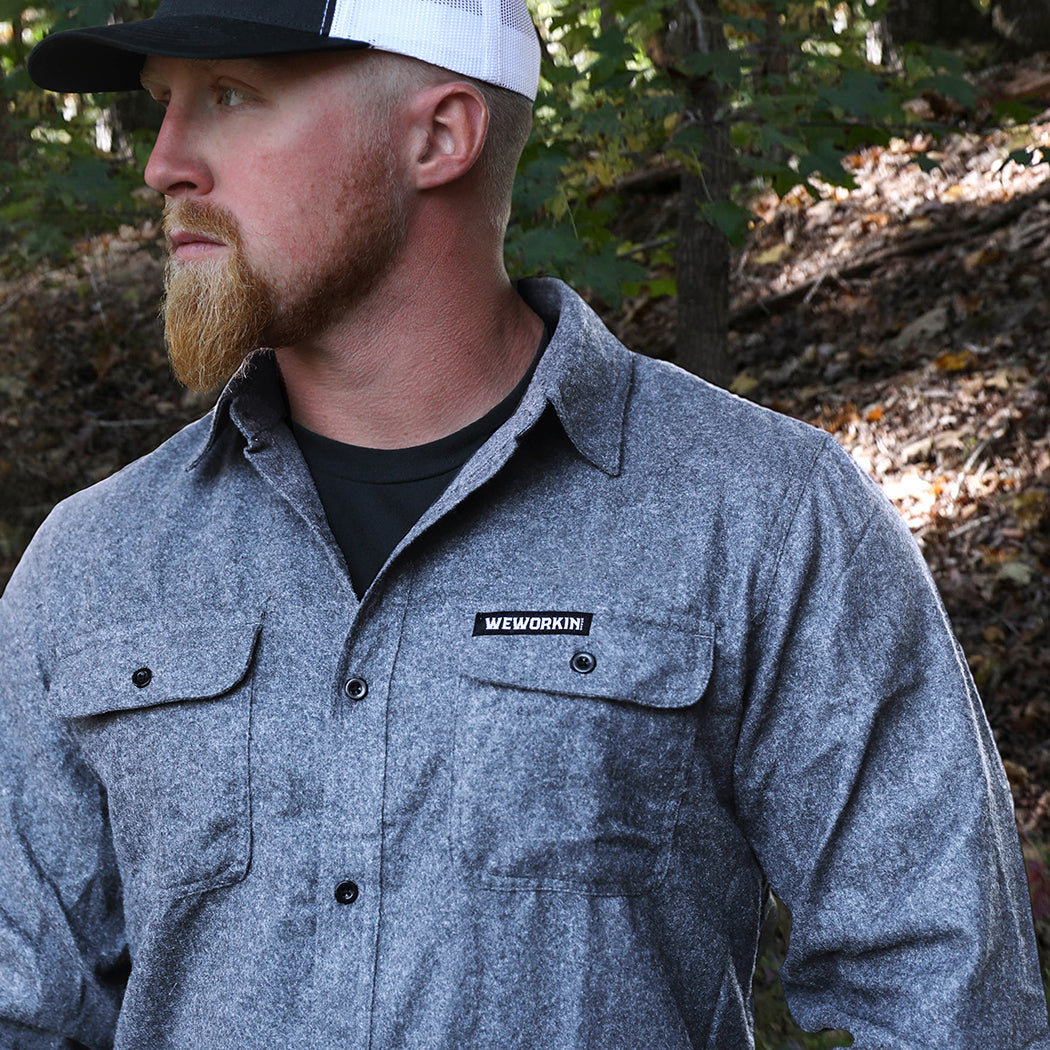 Man pictured from front wearing a We Workin long sleeve Flannel buttoned shirt in Light Heather Grey, wooded background. A small/rectangular black with white thread WEWORKIN BRAND woven tag is sewn on the left pocket flap. He is also wearing a We Workin Script Retro Trucker hat in Black/White.