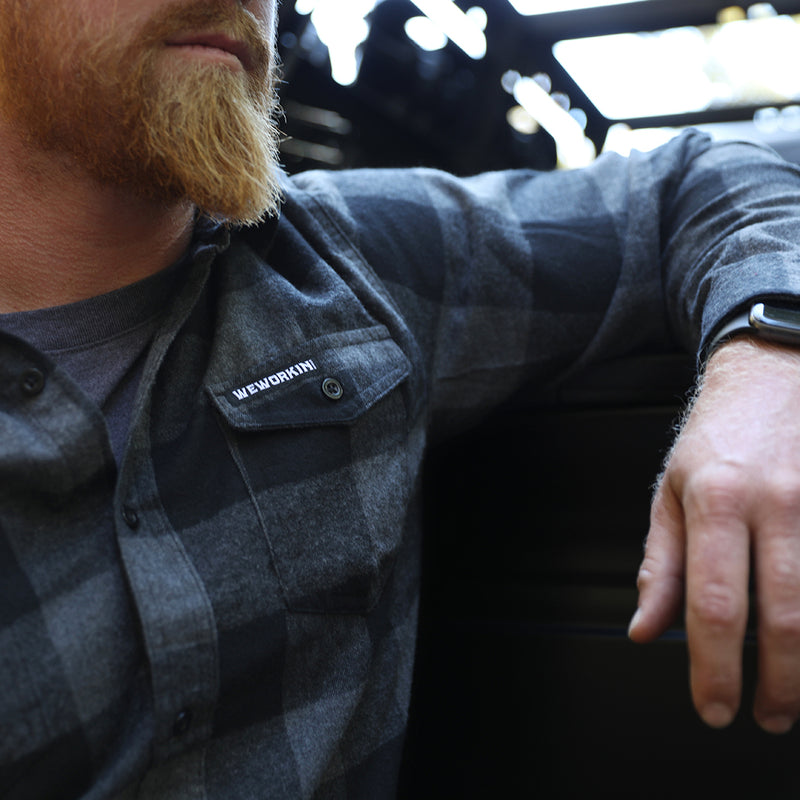 Man pictured from front, close up, wearing a We Workin long sleeve Flannel buttoned shirt in Grey/Black Buffalo Plaid. A small/rectangular black with white thread WEWORKIN BRAND woven tag is sewn on the left pocket flap. 