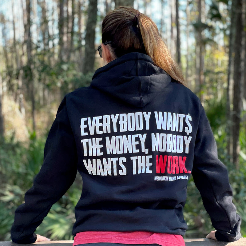 Woman pictured from back wearing a WW HARD Black Hoodie. "EVERYBODY WANT$ THE MONEY, NOBODY WANTS THE WORK" printed in bold on full back width in white ink, except the word WORK is printed in red for emphasis. (WEWORKIN BRAND APPAREL printed small and lower right just below the full back imprint.) Banded cuffs and waist. Hood down on back.