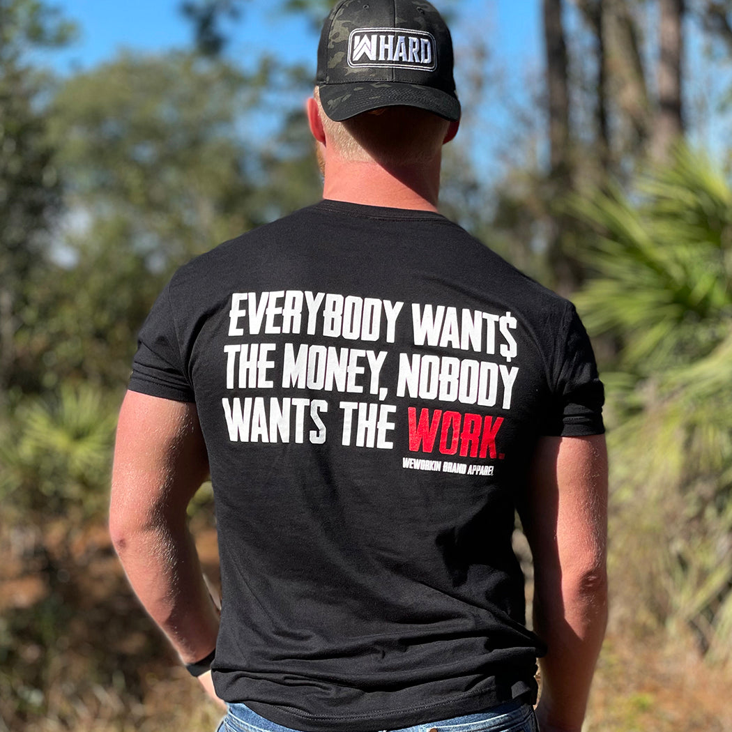 Man pictured from back wearing a WW Black Graphic Tee. "EVERYBODY WANT$ THE MONEY, NOBODY WANTS THE WORK" printed in bold on full back width in white ink, except the word WORK is printed in red for emphasis. (WEWORKIN BRAND APPAREL printed small and lower right just below the full back imprint.) Also wearing a WW HARD Black Camo flexfit hat backwards.