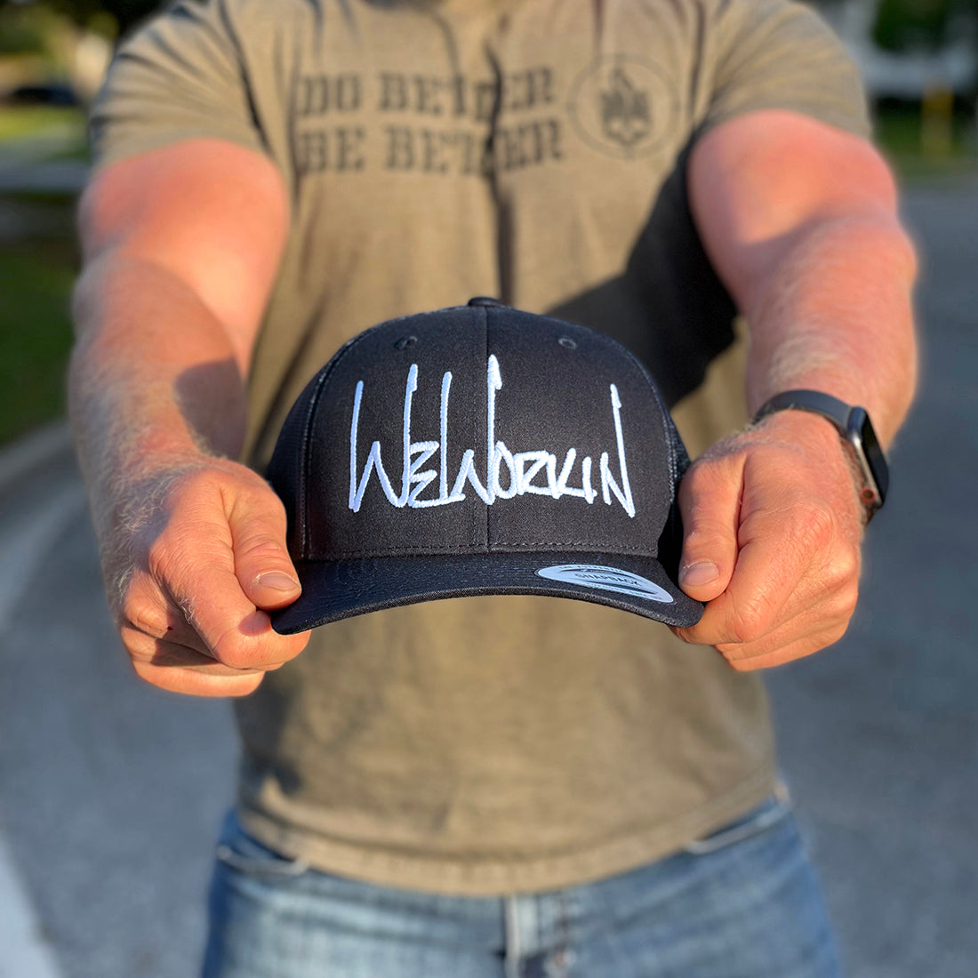 Man holding a WeWorkin Retro Trucker Hat in stealth black color (all panels in black). WeWorkin script logo is embroidered large on the front Black panels in White thread.