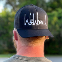 Man wearing a WeWorkin Retro Trucker Hat in Stealth Black. WeWorkin script logo is embroidered large on the front Black panels in White thread.