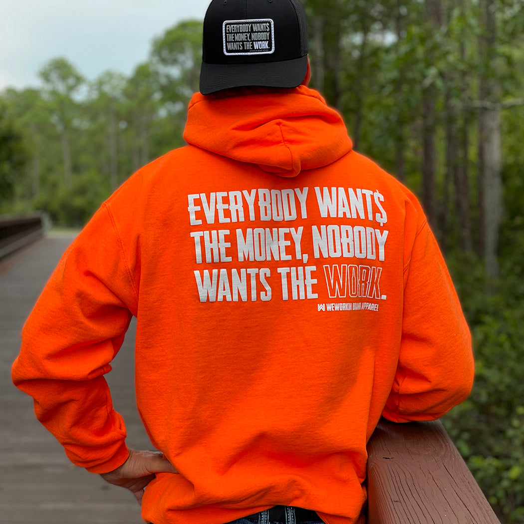 Man pictured from back wearing a We Workin premium Safety Orange hoodie. "EVERYBODY WANT$ THE MONEY, NOBODY WANTS THE WORK" printed large on the back in white ink, and the word WORK is outlined in white for emphasis. (WEWORKIN BRAND APPAREL printed small and lower right just below the full back imprint.) He is also wearing an all black Retro Snapback Patch Hat with same tagline.