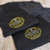 Front "pocket area" of 2 men's short sleeved t-shirts in Black, folded on a tile background. "HARD LIVIN. HARD WORKIN. PROUD AMERICAN." design is printed small, pocket-sized, in gold and grey ink.