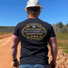 Man pictured from back outdoors, wearing a We Workin Black Graphic Tee. "HARD LIVIN. HARD WORKIN. PROUD AMERICAN." design is printed in bold on full back width in gold and grey ink.