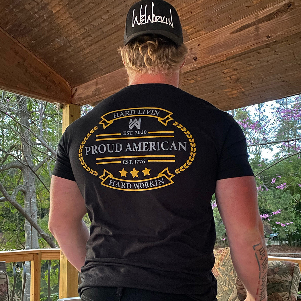 Man pictured from back wearing a We Workin Black Graphic Tee. "HARD LIVIN. HARD WORKIN. PROUD AMERICAN." design is printed in bold on full back width in gold and grey ink. Also wearing a WEWORKIN Script logo (white embroidery) Retro Trucker hat backwards.