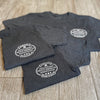 Front "pocket area" of 3 men's short sleeved t-shirts in Charcoal Grey color, folded on a tile background. "HARD LIVIN. HARD WORKIN. PROUD AMERICAN." design is printed small, pocket-sized, in white ink.