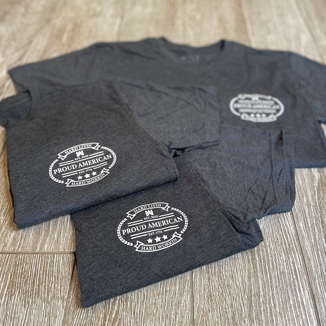 Front "pocket area" of 3 men's short sleeved t-shirts in Charcoal Grey color, folded on a tile background. "HARD LIVIN. HARD WORKIN. PROUD AMERICAN." design is printed small, pocket-sized, in white ink.