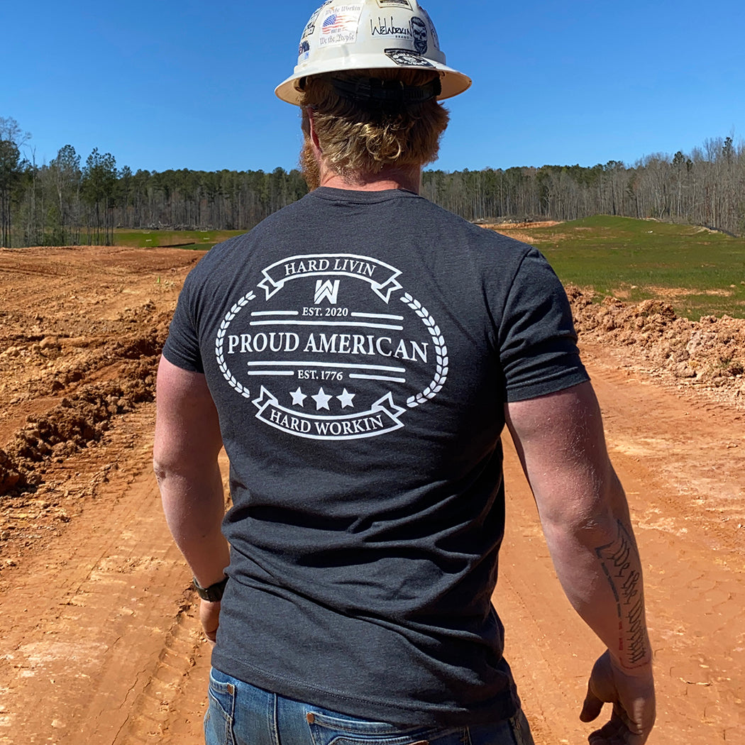 Man pictured from back, outdoors, wearing a We Workin Charcoal Grey Graphic Tee. "HARD LIVIN. HARD WORKIN. PROUD AMERICAN." design is printed in bold on full back width in white ink.