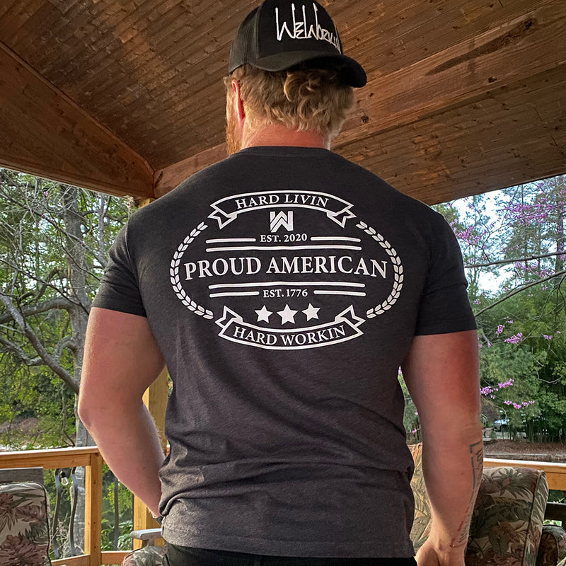 Man pictured from back wearing a We Workin Charcoal Grey Graphic Tee. "HARD LIVIN. HARD WORKIN. PROUD AMERICAN." design is printed in bold on full back width in white ink. Also wearing a WEWORKIN Script logo (white embroidery) Retro Trucker hat backwards.