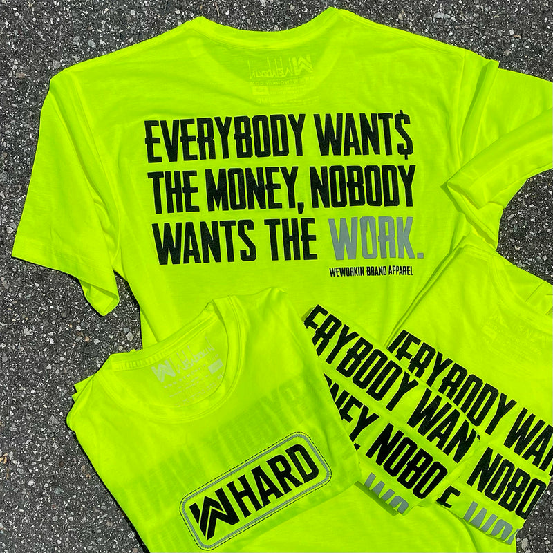 We Workin Hi-Viz short sleeve tees in Safety Yellow laying on an asphalt road. Back is printed with the tagline "Everybody Wants the Money, Nobody Wants the WORK." in black ink, except the word WORK is in grey ink. Front of tee is printed with the We Workin WWHARD patch-style icon printed in Black ink, except the "rope" style outline is in grey ink.