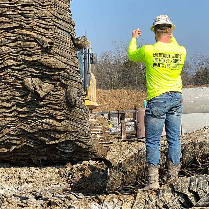 Man on construction site, pictured from back, wearing the We Workin Hi-Viz Safety Yellow long sleeve tee. Back is printed with the tagline "Everybody Wants the Money, Nobody Wants the WORK." in black ink, except the word WORK is in grey ink.