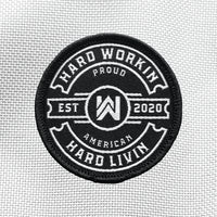 "HARD WORKIN. HARD LIVIN." text circling the WW icon logo and "EST 2020"  text in center, on a velcro-backed patch (both the hook and loop velcro sides included). [1] thread color for the design (white) on a black woven background, with black merrowed border. "PROUD AMERICAN" circles top and bottom under outer text. 2.25" wide circular Woven patch displayed on a grey canvas background.