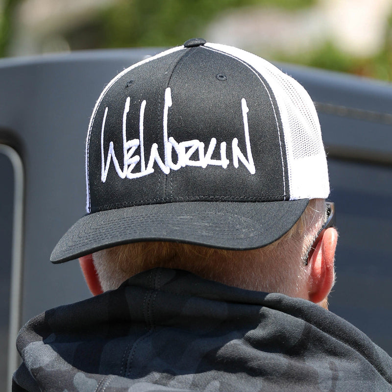 Man wearing a WeWorkin Retro Trucker Hat in Black/White. WeWorkin script logo is embroidered large on the front Black panels in White thread.