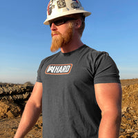 Man viewed from the front wearing a WW HARD Tee in Heather Charcoal Grey. WW HARD "patch-style" icon printed in Neon Orange and White, small on center chest (pictured on a jobsite with a hardhat on—WeWorkin Hard Hat stickers on hat)