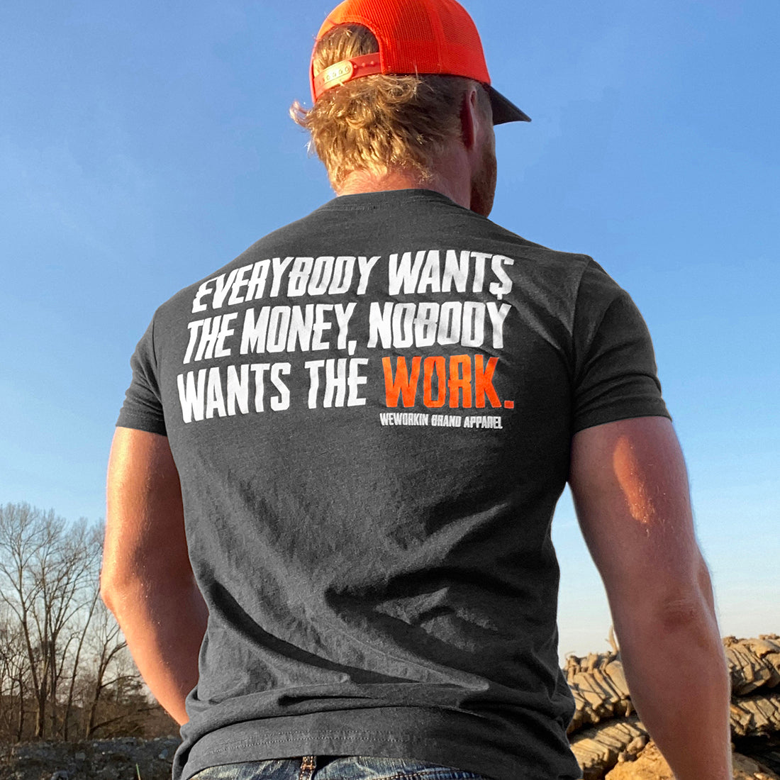 Man pictured from back wearing a WW Charcoal Heather Grey Graphic Tee. "EVERYBODY WANT$ THE MONEY, NOBODY WANTS THE WORK" printed in bold on full back width in white ink, except the word WORK is printed in Neon Orange for emphasis. (WEWORKIN BRAND APPAREL printed small and lower right just below the full back imprint.) Also wearing a WW HARD Blaze Orange/Grey Retro Trucker Hat.