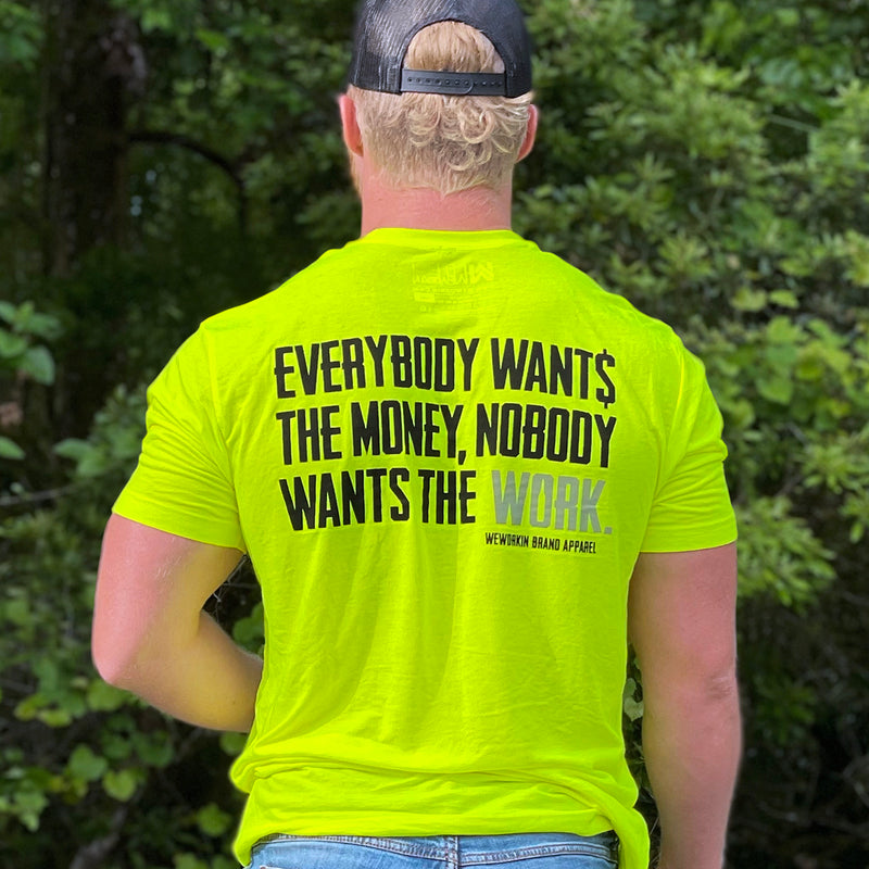 Man outdoors, pictured from back, wearing the We Workin Hi-Viz Safety Yellow short sleeve tee. Back is printed with the tagline "Everybody Wants the Money, Nobody Wants the WORK." in black ink, except the word WORK is in grey ink—and the stealth black We Workin Snapback.