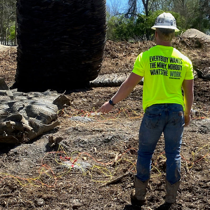 Man on construction site, pictured from back, wearing the We Workin Hi-Viz Safety Yellow short sleeve tee. Back is printed with the tagline "Everybody Wants the Money, Nobody Wants the WORK." in black ink, except the word WORK is in grey ink.