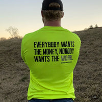 Man outdoors, pictured from back, wearing the We Workin Hi-Viz Safety Yellow long sleeve tee. Back is printed with the tagline "Everybody Wants the Money, Nobody Wants the WORK." in black ink, except the word WORK is in grey ink.