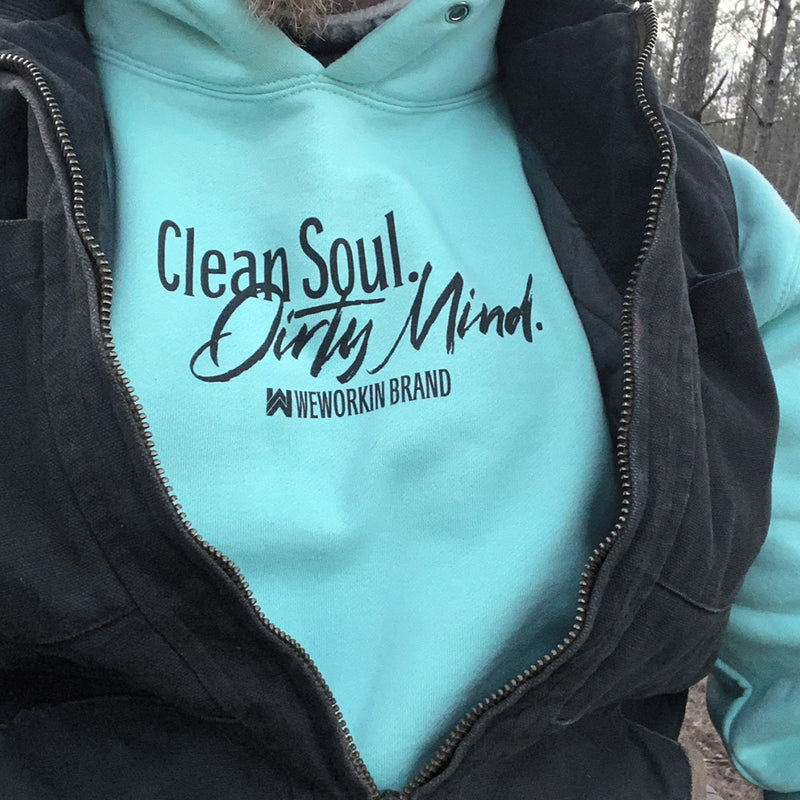 Close up, front view of man wearing the medium-weight mint teal long-sleeve hooded sweatshirt (under a black canvas vest). View is cropped in close on the "Clean Soul. Dirty Mind." tagline printed on center chest (WW icon and WEWORKIN BRAND text under tagline, small and centered.) Thick drawstring in hood, nickel eyelets.