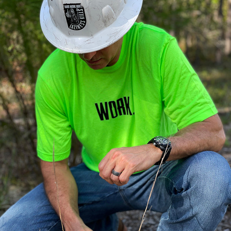 Man outdoors with a hard hat on kneeling down, wearing the We Workin Hi-Viz short sleeve tee in Safety Green color. Front is printed with the "WORK." in black ink. The letter "O" in WORK has a bullet in the center to make the inside of the "O".