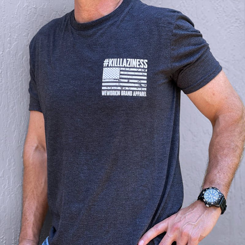 Man pictured from the front wearing a Steel Grey WeWorkin short sleeve tee. Graphic printed in place of a left check pocket. Graphic includes a bold grunge-style US flag and the text #KILLLAZINESS above, and WEWORKIN BRAND APPAREL below the flag, all printed in white ink. 