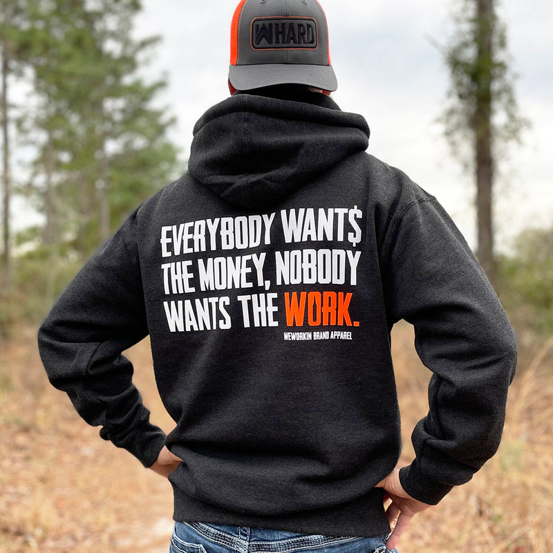 Man pictured from back wearing a WW HARD Charcoal Grey Hoodie. "EVERYBODY WANT$ THE MONEY, NOBODY WANTS THE WORK" printed in bold on full back width in white ink, except the word WORK is printed in neon orange for emphasis. (WEWORKIN BRAND APPAREL printed small and lower right just below the full back imprint.) Banded cuffs and waist. Hood down on back. (Also wearing a WW HARD Grey/Neon Orange Retro Trucker hat backwards.)